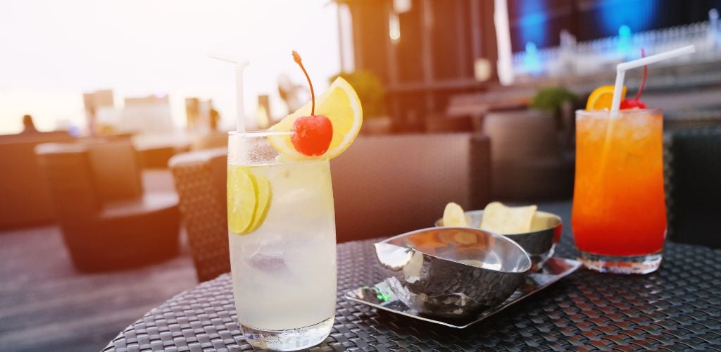 Two glasses of cocktails, one with lemon and cherry, and the other with orange and cherry, in an outdoor ambiance. In addition, it has served two metal bowls.