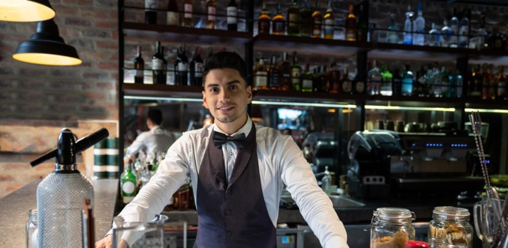 A male bartender is standing on the bar corner with a lot of wine glasses in the background.