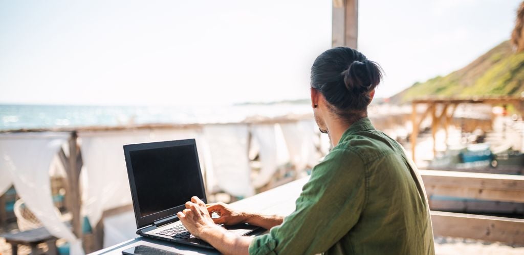 digital nomad working on a laptop in a beach cafe location overlooking sunbeds and the ocean 