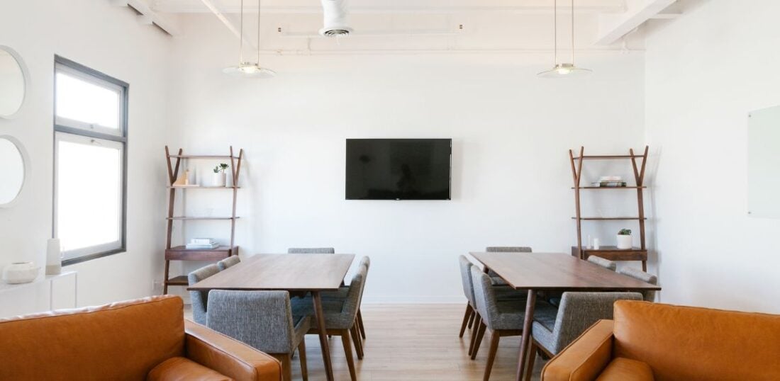 office space with comfortable couch style seating and a TV screen mounted on the wall. Two individual desks in background of image