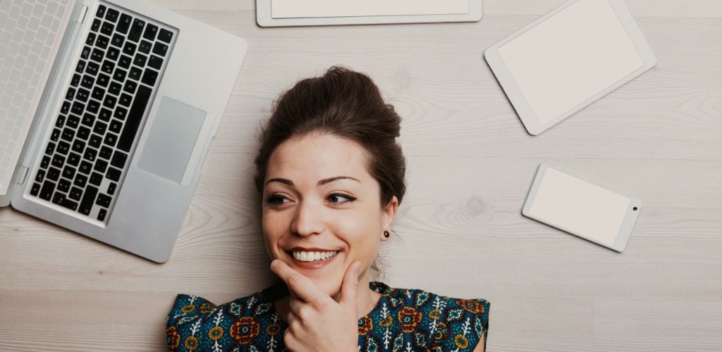 woman laying on floor thinking with smile on face surrounded by laptop and tablet and mobile 