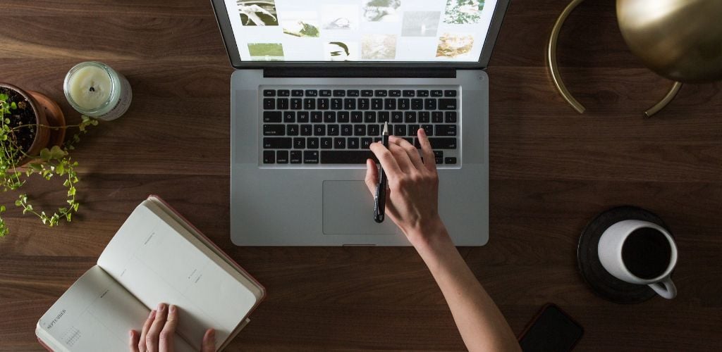 person completing research from laptop, looking at images on screen whilst taking notes, coffee mug, candle, and plant on desk 