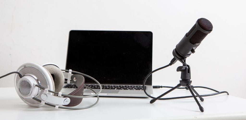 podcast set up on white desk with white wall behind. Laptop, headphones, and microphone