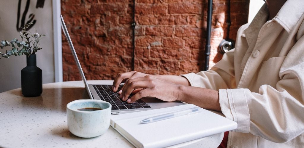 person typing on a laptop with a coffee and a notebook to side, exposed brick wall in background