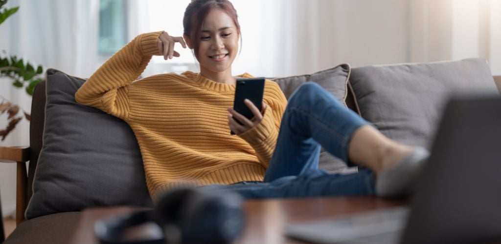 woman smiling at phone with laptop close by sitting on sofa 