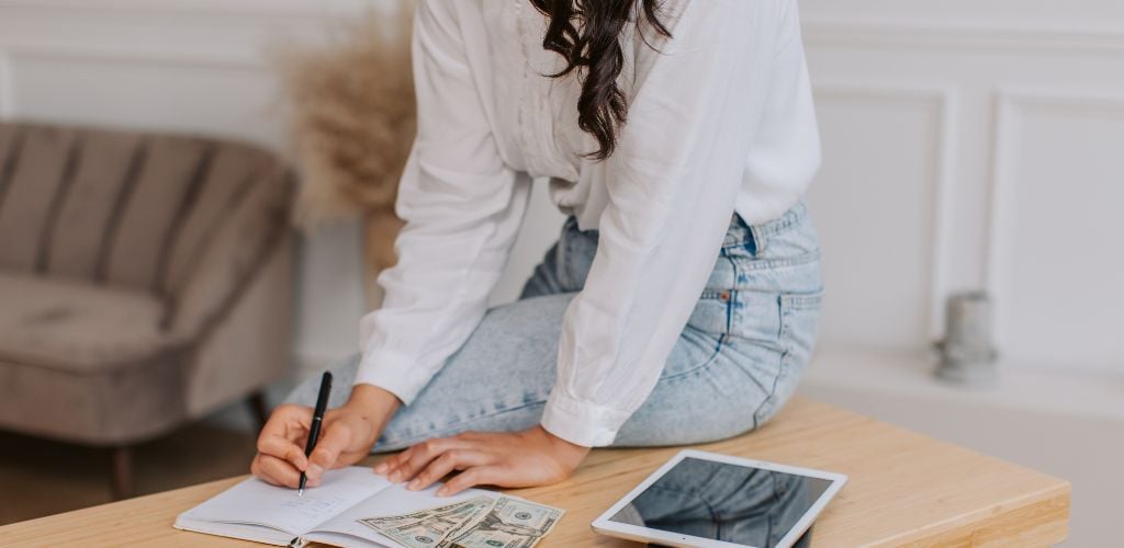 woman sitting on desk working out budget, dollars on notebook and tablet device to the side on desk 