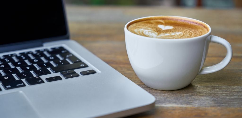 image of laptop on cafe table with latte mug beside keyboard, latte heart features a heart and leaf pattern 