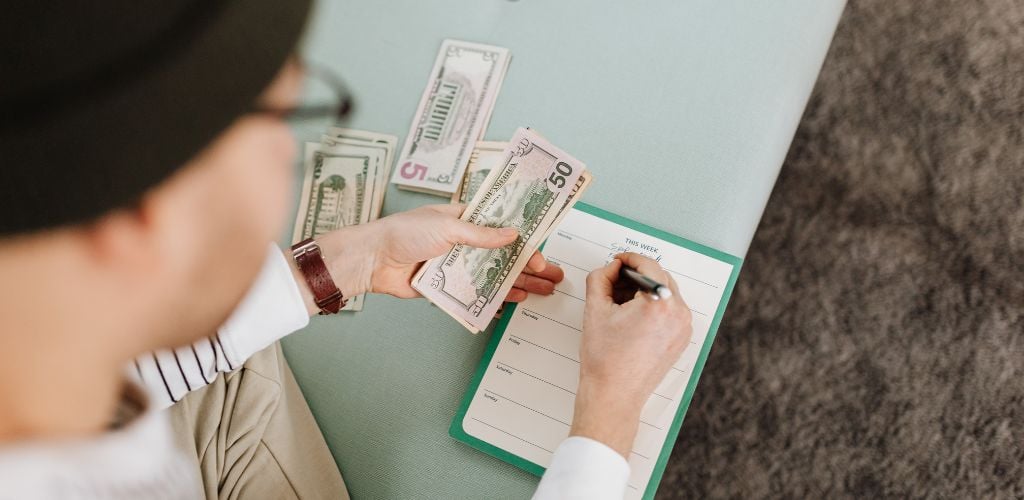 man working out weekly budget, writing on notepad whilst holding dollars in other hand 