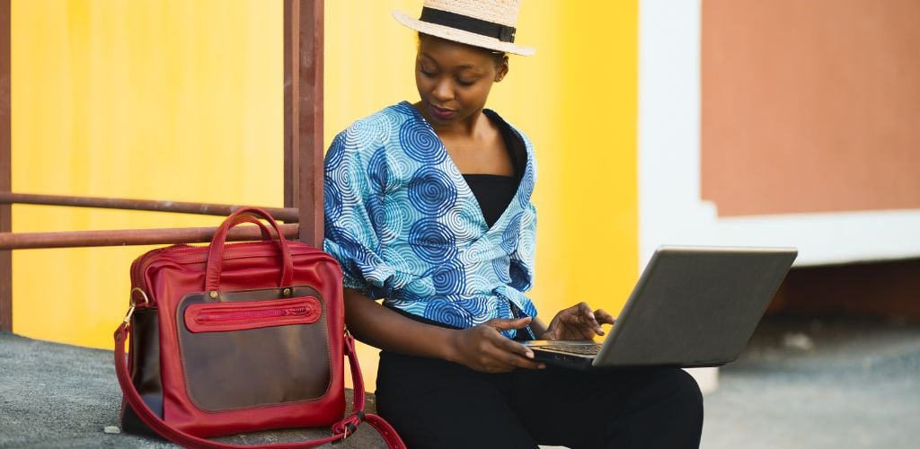 woman sitting outdoors working remotely from laptop with laptop bag beside her 