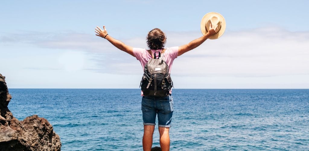 male traveller standing in front of horizon viewpoint with arms reaching out and hat in hand