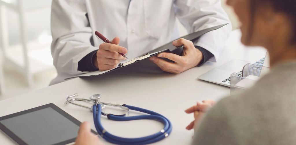 medical professional in white coat filling out form with patient on opposite side of desk, iPad and stethoscope on desk