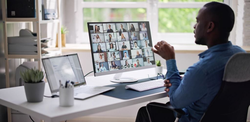 man sitting in home office environment, with online meeting in progress. Multiple attendees visible on computer screen. 