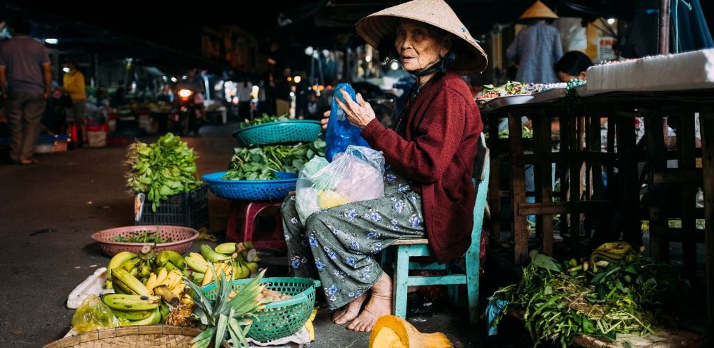 A market vendor sitting amongst fresh local produce such as figs, bananas, local greens and pineapple