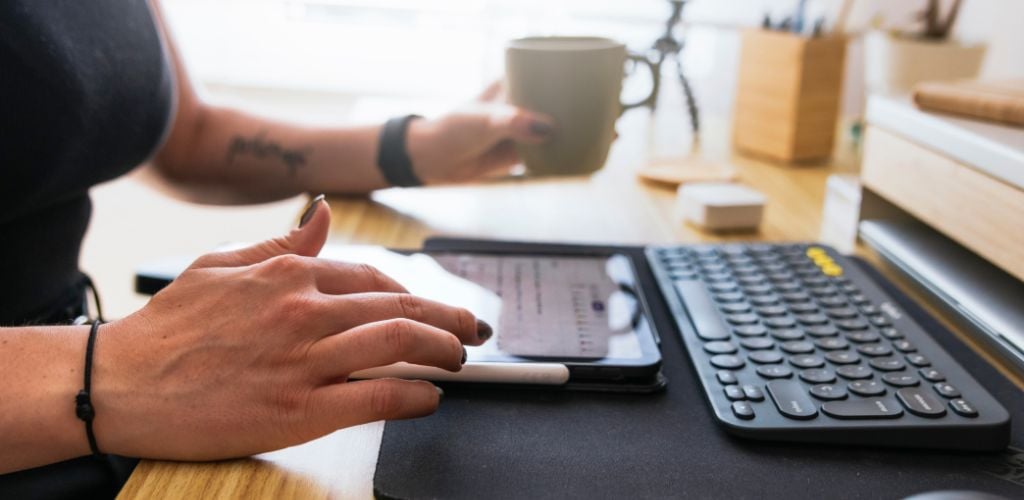 person using tablet device, coffee mug in hand at desk with keyboard 