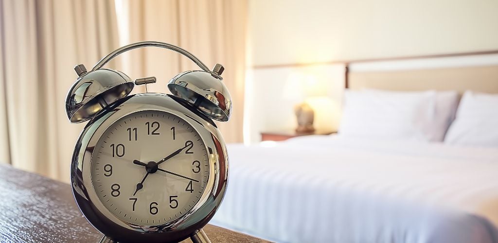 Traditional alarm clock on wooden table with bed in background. Clock shows 07:10 am. 