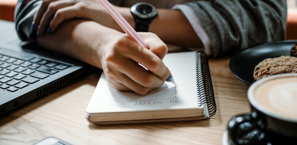 person writing a to-do list, laptop to the side and coffee and cookies on desk 