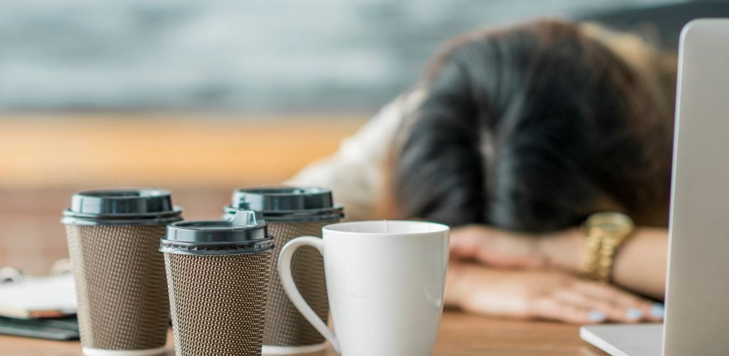 woman asleep on desk, multiple take-out coffee cups on desk with laptop open 