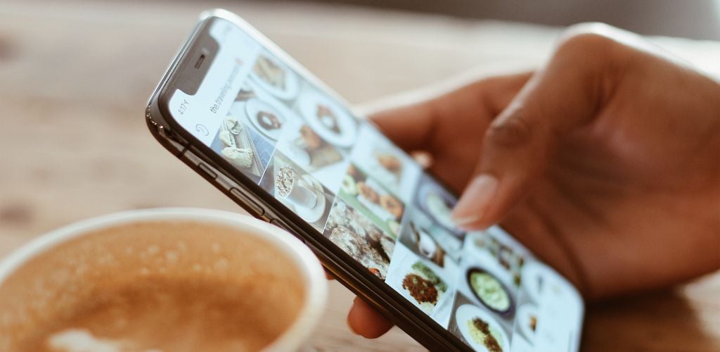 person holding smartphone in hand with Instagram social media page on screen, coffee mug on table