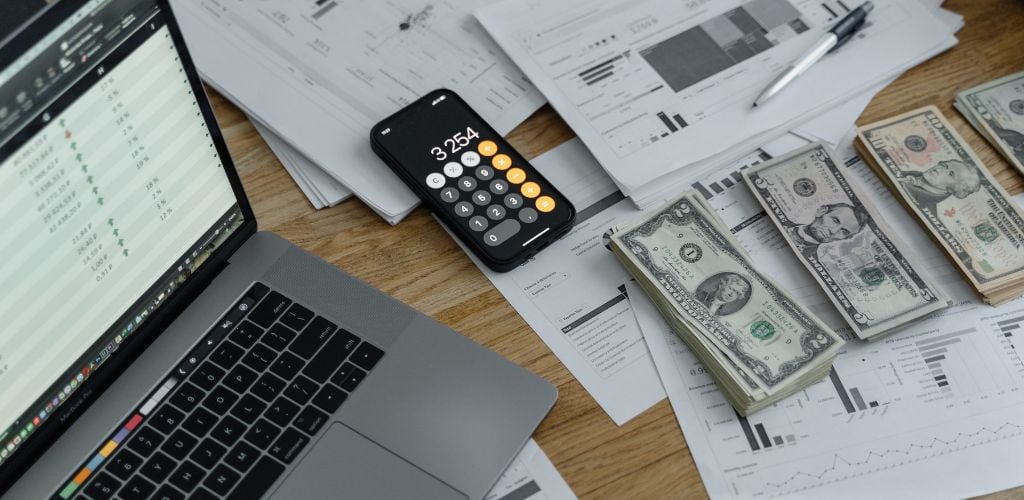 laptop open on desk with spreadsheet and stocks displayed, mobile phone with calculator open, printed spreadsheets on paper, and piles of organised dollars 