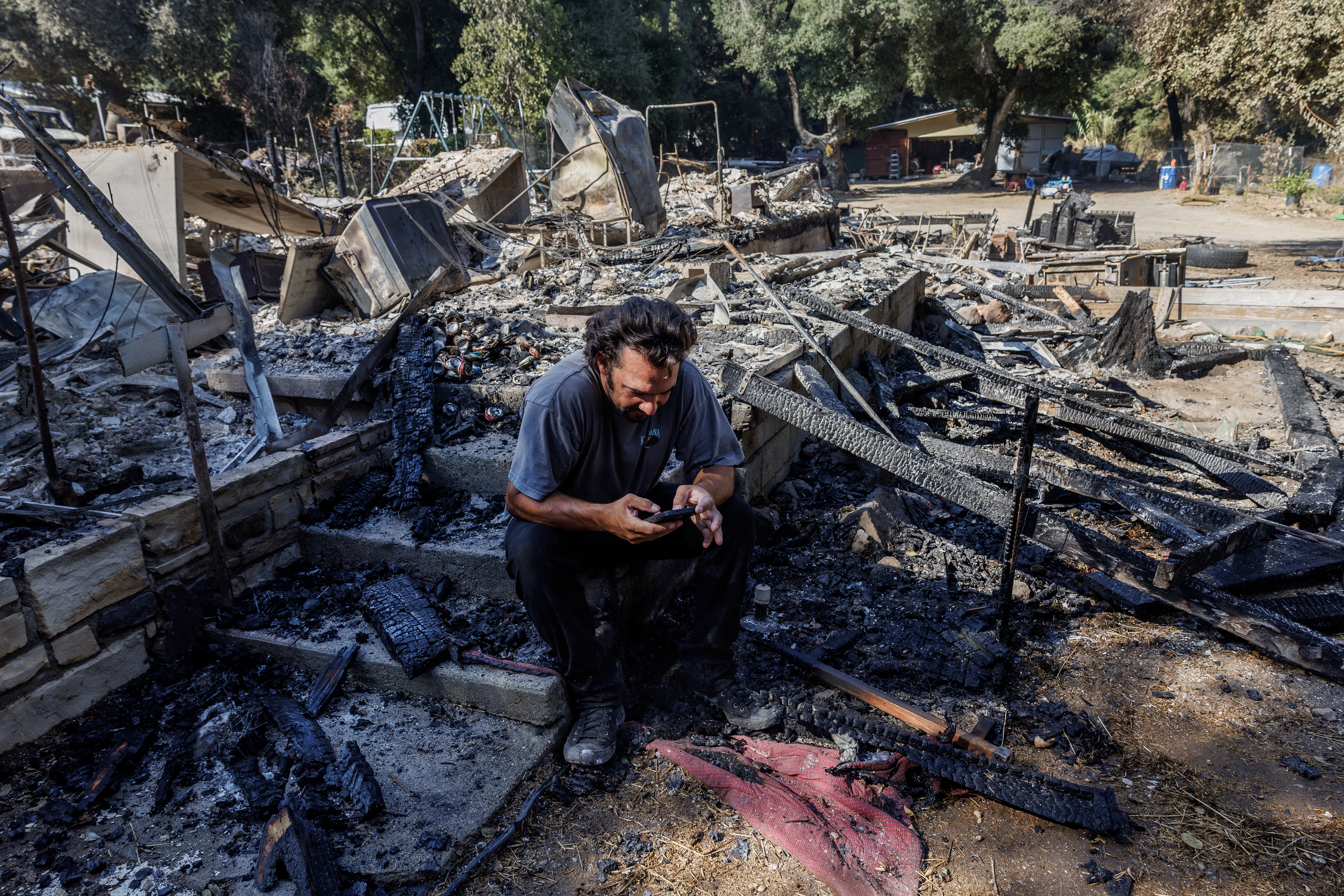 Garrett Keene sits on the front steps of his home in Lake Elsinore, Calif. that was destroyed in the Airport Fire, one of three major Southern California Fires so far this September. After evacuating with his family, Keene said he monitored the Watch Duty fire app to see where airdrops of fire retardant were happening as the fire approached, but none dropped on his neighborhood.