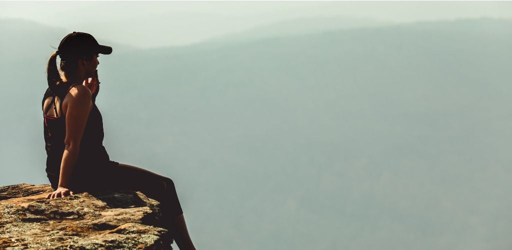 woman sitting at edge of cliff after hike, wearing workout gear and cap 