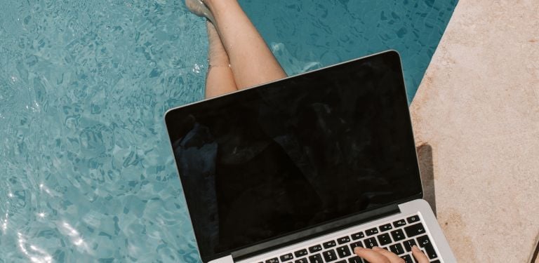 digital nomad working on laptop whilst dipping legs into a pool 